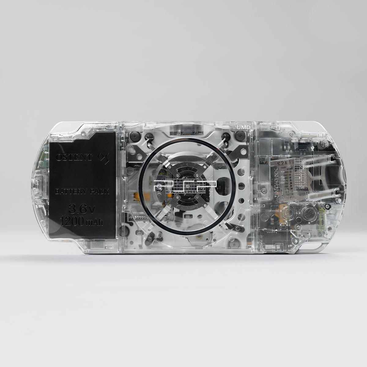 Back view of a transparent Sony PSP with grey buttons, showing internal UMD drive, OSTENT 3.6V 1200mAh battery pack, and clear shell design