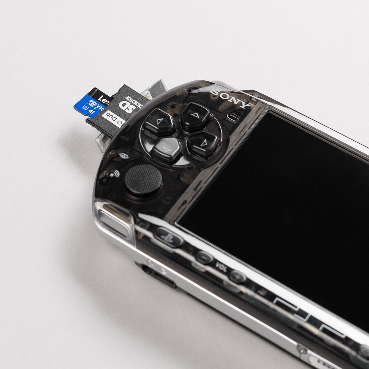 Close-up of a black transparent PSP with a dual-slot microSD to Memory Stick Duo adapter inserted, showing clear shell and black button mod