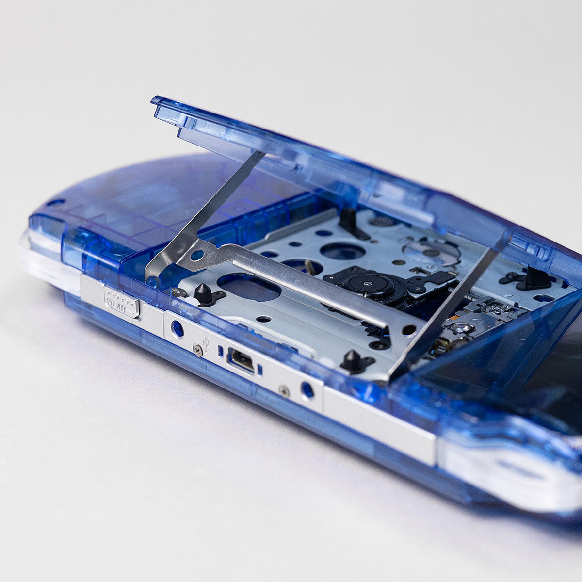 Detailed view of a blue transparent PSP with the UMD drive door open, showing clean internal disc tray and laser mechanism beneath the shell