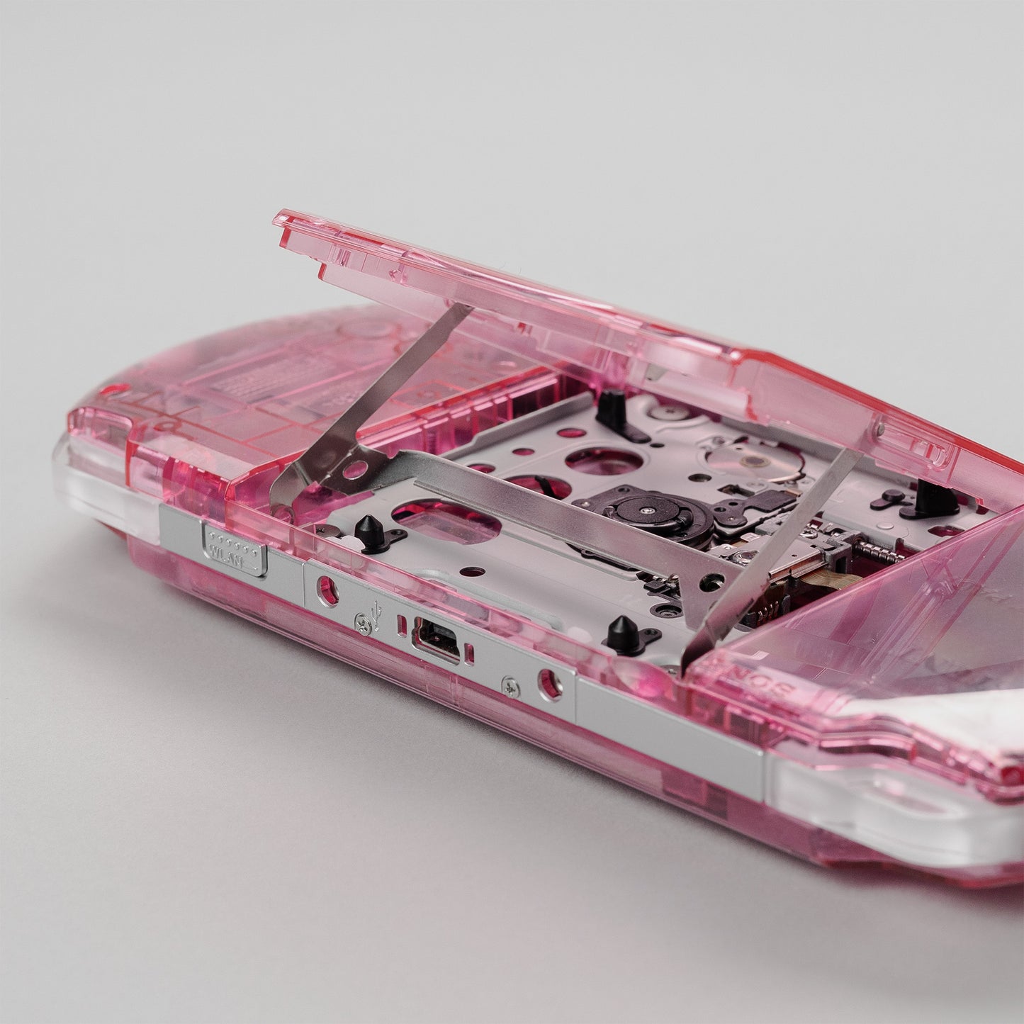 Close-up of a pink transparent PSP with the UMD drive door open, showing the internal disc tray and laser assembly in detail