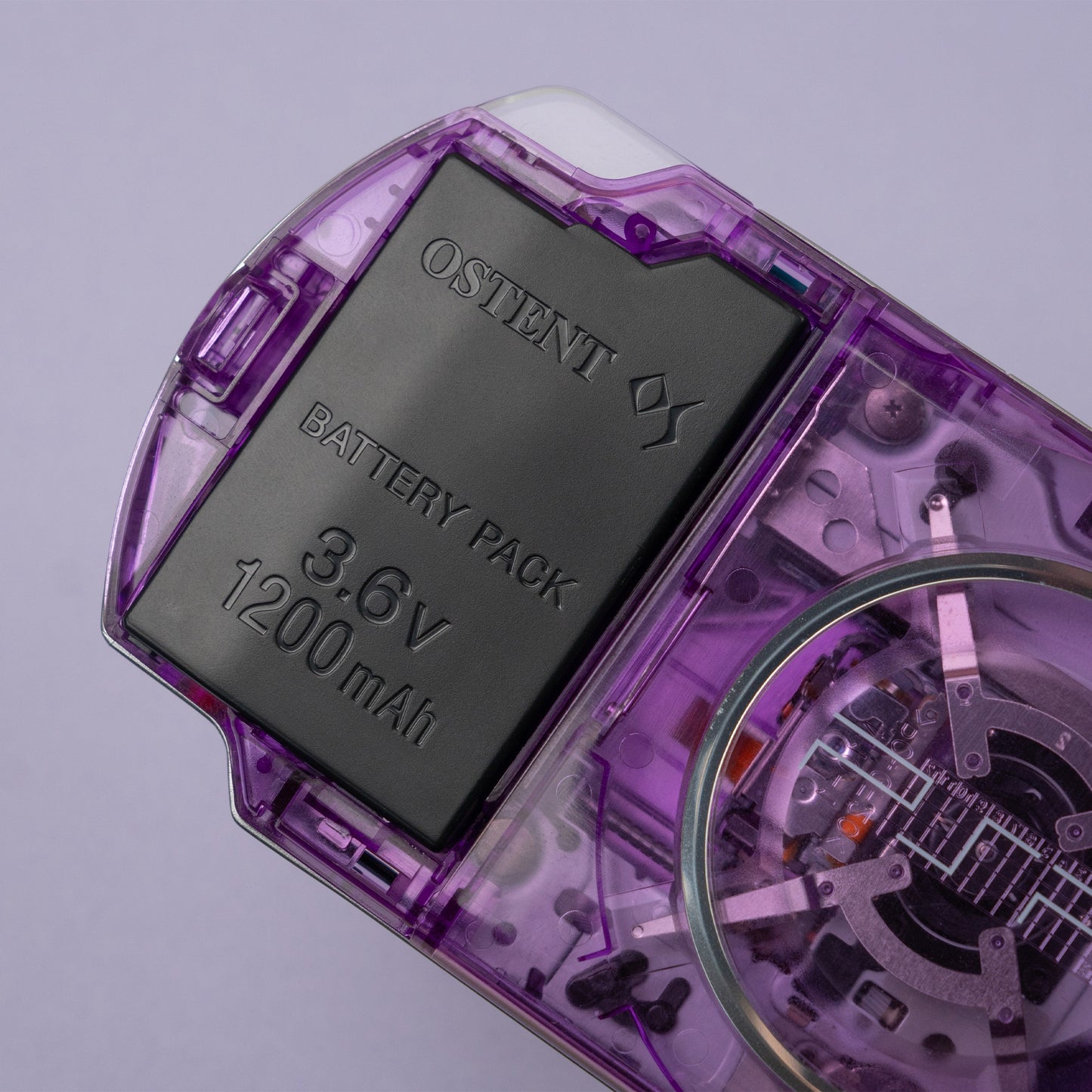 Close-up of a purple transparent PSP showing OSTENT 3.6V 1200mAh battery pack and UMD drive internals through the clear shell design