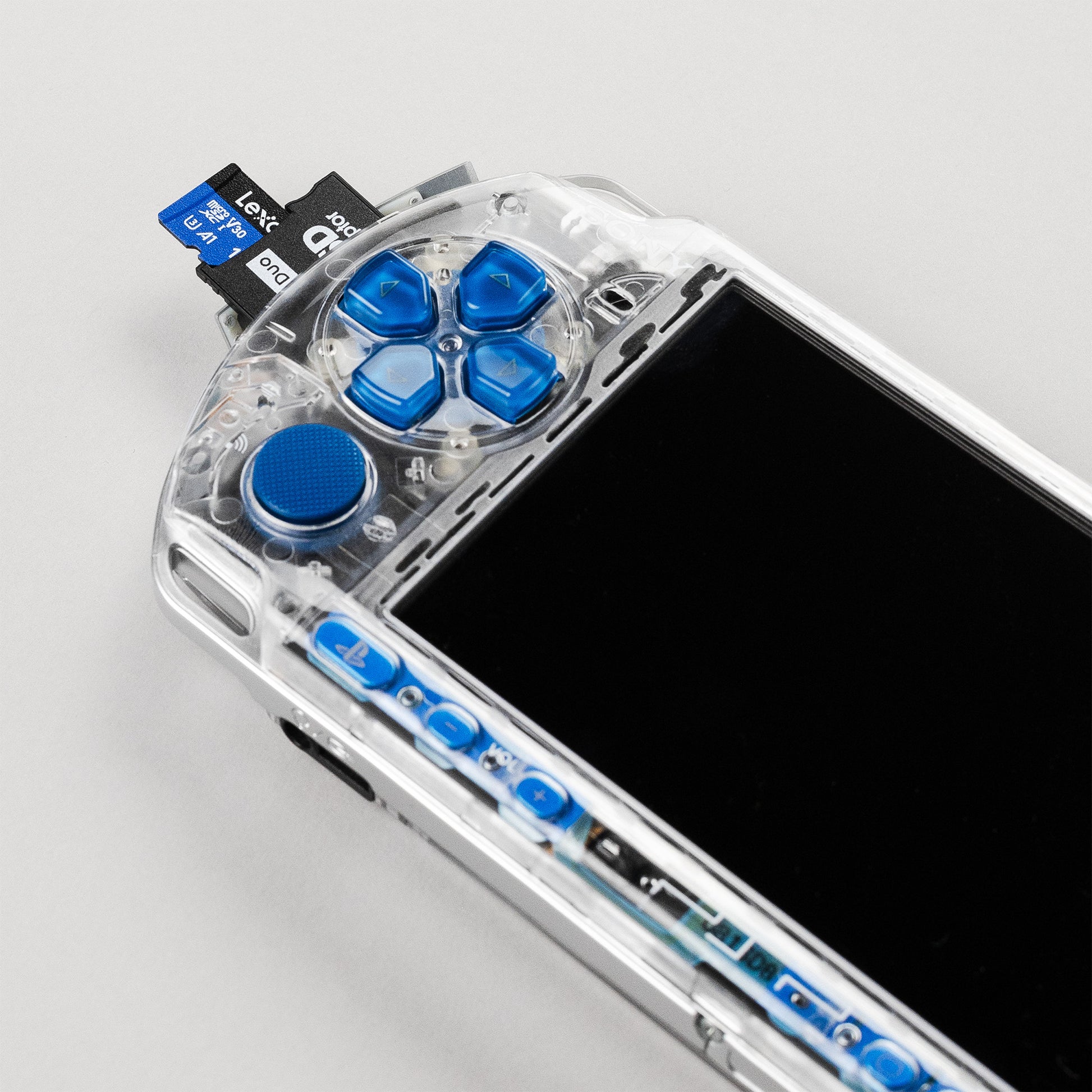 Close-up of a clear PSP with blue buttons and a dual-slot microSD to Memory Stick Pro Duo adapter inserted, showing a Lexar 128GB card