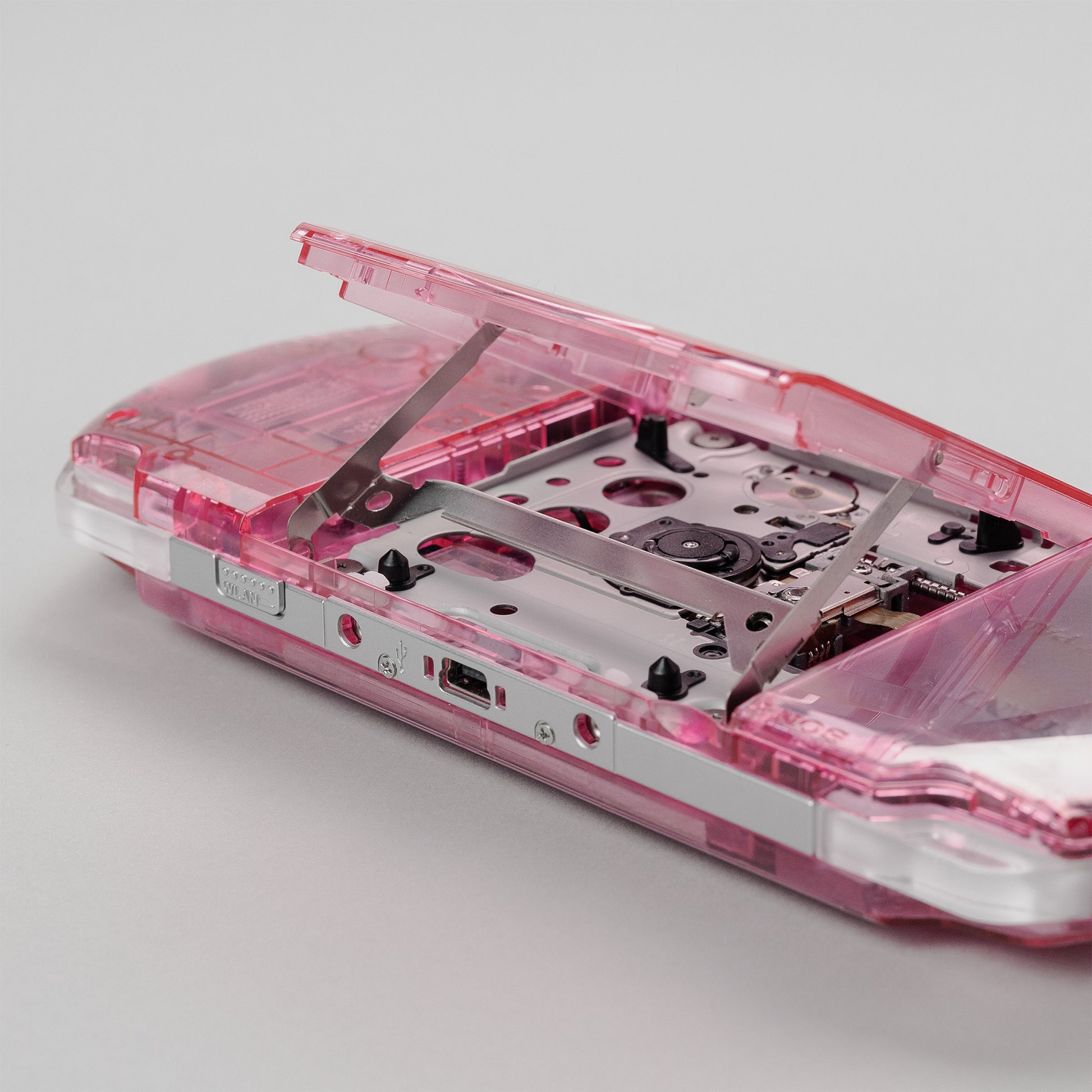Close-up of a pink transparent PSP with the UMD drive door open, showing the internal disc tray and laser assembly in detail