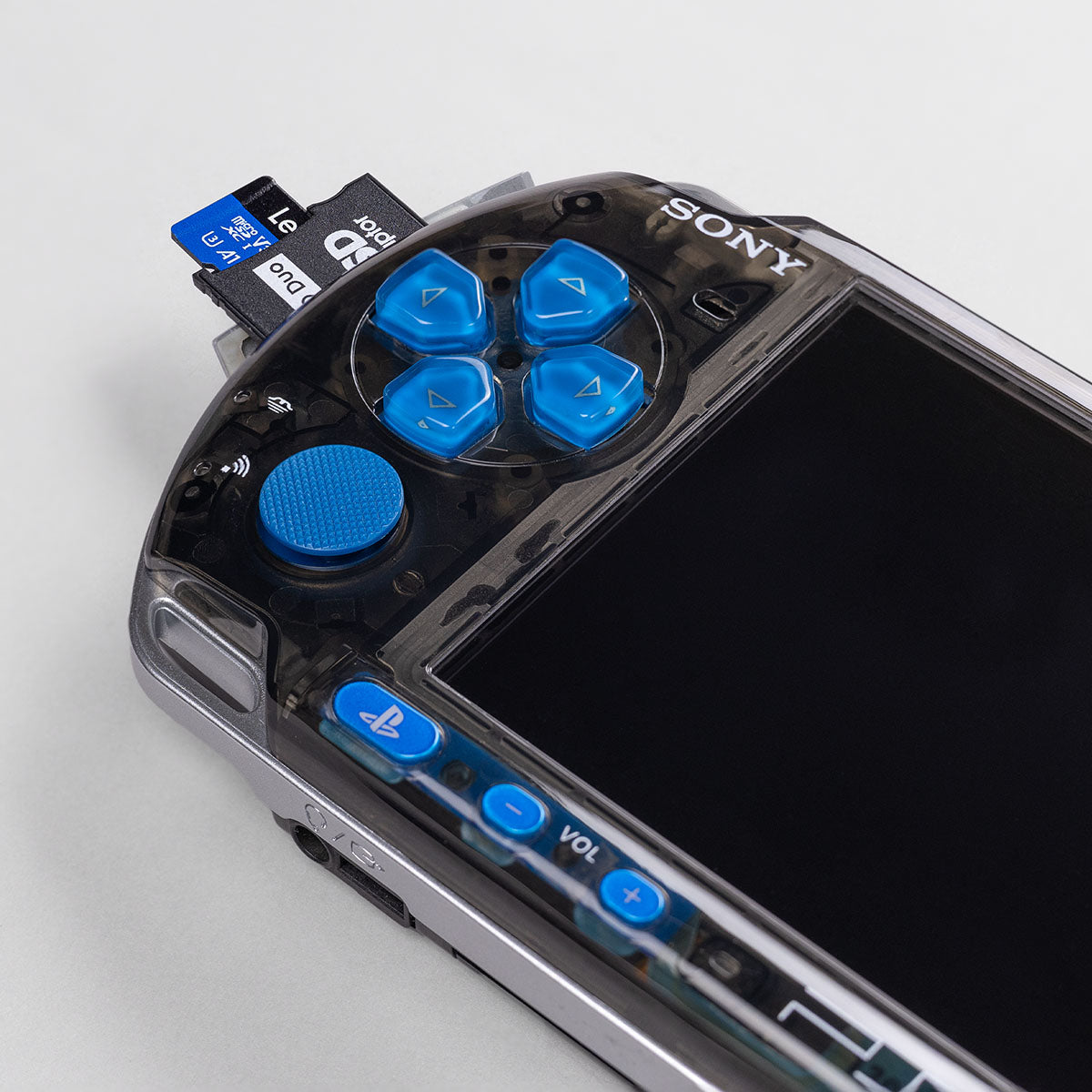 Close-up of a transparent black PSP-3000 with blue buttons and analog stick, showing a dual microSD adapter inserted in the Memory Stick slot.