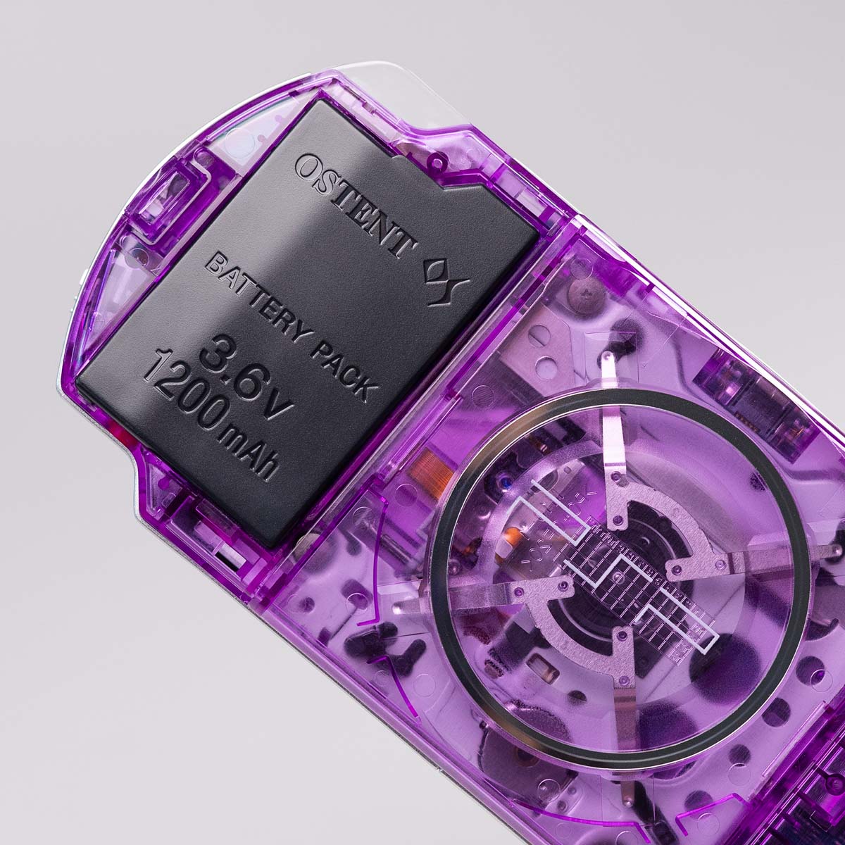 Close-up of a purple transparent PSP showing the OSTENT 3.6V 1200mAh battery pack and internal UMD drive through the clear rear shell