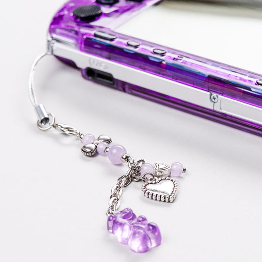 Close-up of a purple Sony PSP with a kawaii charm accessory featuring silver heart beads, lilac pearls, and a translucent purple gummy bear pendant