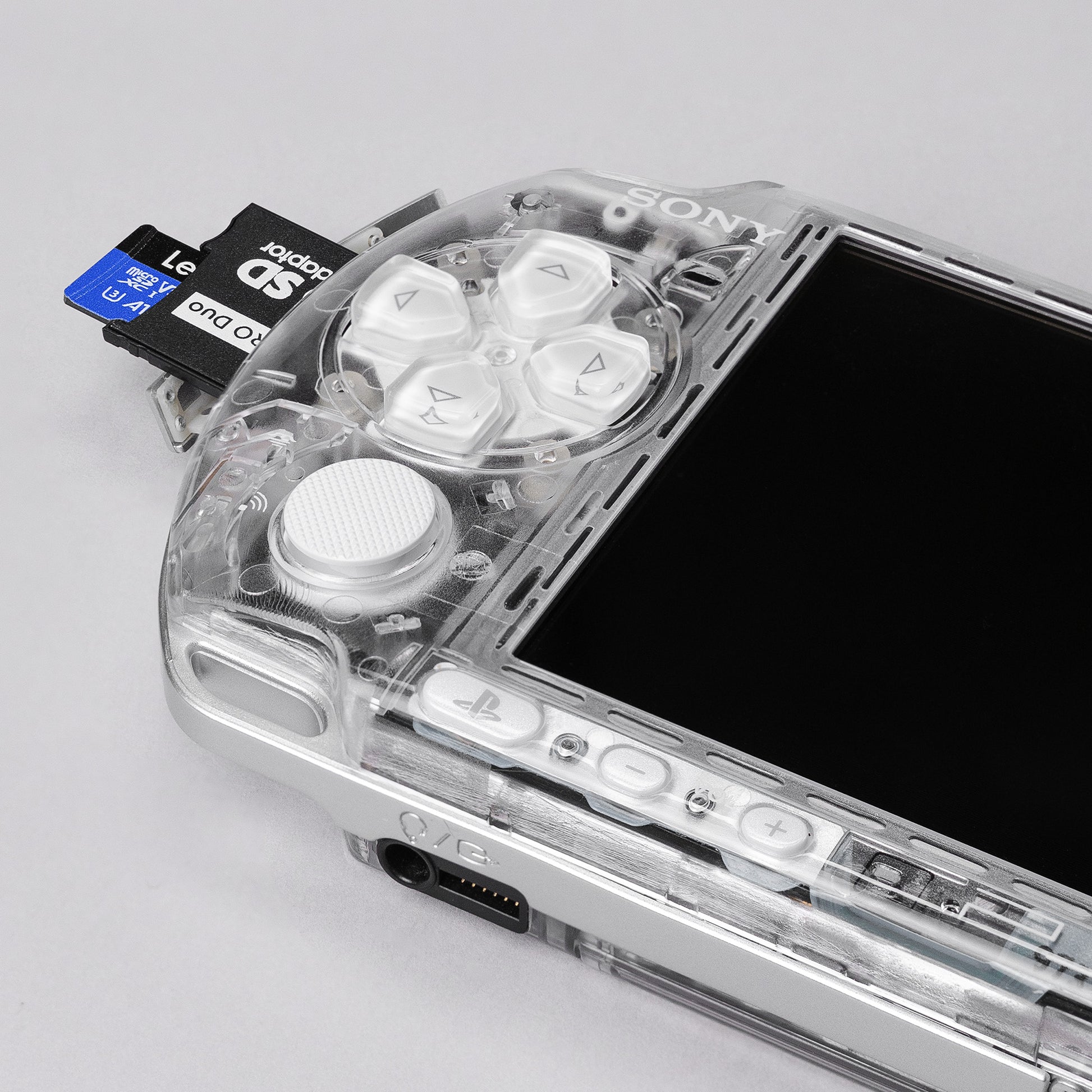 Close-up of a Sony PSP with transparent clear shell and white buttons, showing a dual microSD to Memory Stick Pro Duo adapter inserted